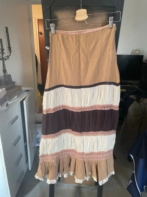 Bohemisk långkjol från Vila - Säljer en bohemisk långkjol från Vila i beige, brun och vit med volangkant nertill och dekorativa spetsdetaljer. Kjolen har ett avslappnat fall. 