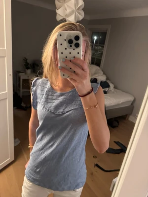 Ljusblå volang blus med spetsdetalj - Supersöt ljusblå blus med virkade spetsdetaljer upptill och ärmlös design. Perfekt till jeans eller kjol för en fräsch look. Blusen har en mjuk och luftig känsla, vilket gör den skön att bära under varmare dagar.