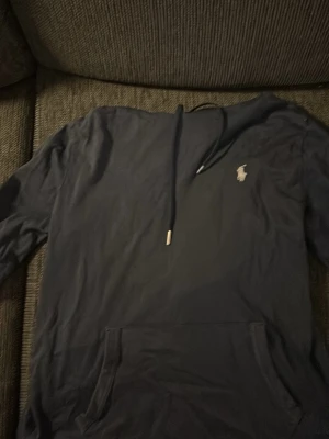 Mörkblå hoodie från Polo Ralph Lauren - Snygg mörkblå hoodie från Polo Ralph Lauren med klassisk logga broderad på bröstet. Hoodien har huva med dragsko och en stor magficka framtill. Tillverkad i mjukt bomullsmaterial som känns skönt mot huden. Perfekt för en avslappnad och stilren look.