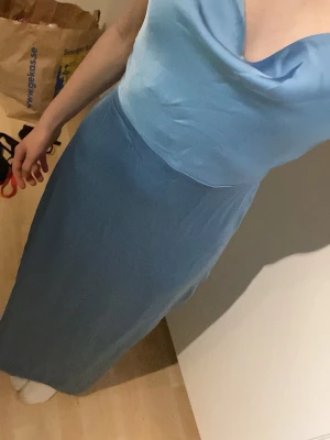 Ljusblå lång klänning med smala band - Säljer en elegant ljusblå lång balklänning med smala axelband och draperad urringning. Klänningen har en figurnära passform och är tillverkad i ett glansigt, mjukt tyg som ger en lyxig känsla. 