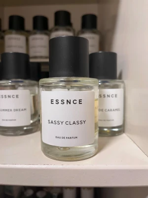 Essnce Sassy Classy Eau de Parfum - Essnce Sassy Classy Eau de Parfum i en rund, transparent glasflaska med svart matt cylinderformad kork. Parfymen har en ljusgul nyans och minimalistisk vit etikett med svart text. Perfekt för dig som gillar stilrena och moderna dofter.