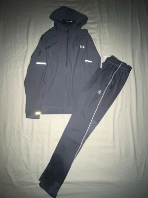 Svart träningsset från Under Armour - Snyggt svart träningsset från Under Armour med huva på jackan och dragkedja framtill. Setet har vita detaljer och reflexband på både jacka och byxor. Perfekt för löpning eller gym. Materialet är lätt och andas bra.