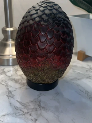Drakägg prydnad Game of Thrones - Säljer en cool drakäggs-prydnad inspirerad av Game of Thrones. Ägget har en skrovlig yta med fjällmönster i svart och mörkrött, samt en grönaktig botten. Perfekt som dekoration för fantasyfans eller som unik inredningsdetalj.