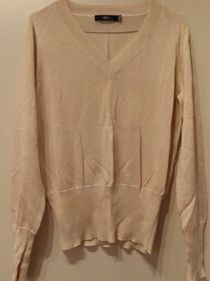 Beige stickad tröja från Ellos - Snygg beige stickad tröja från Ellos med V-ringning och långa ärmar. Tröjan har en lätt glittrig finish och ribbade muddar vid ärmslut och nederkant. Perfekt för dig som gillar en enkel men ändå elegant look.