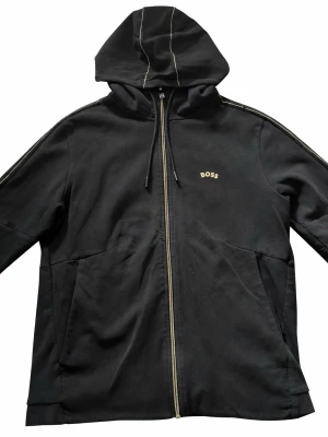 Svart hoodie med dragkedja från BOSS - Snygg svart hoodie från BOSS med dragkedja framtill och diskret BOSS-logga på bröstet. Hoodien har guldiga kontrastsömmar på huvan och axlarna samt praktiska fickor på sidorna. Tillverkad i mjukt bomullsmaterial och har en avslappnad passform.