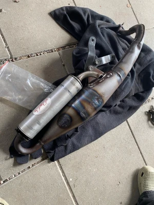 Leovince tt rör - Säljer ett avgassystem i metall med böjd form, troligen till moped eller mindre motorcykel. Färgen är mestadels metallisk med inslag av blått och brunt från värme. Monteringsfästen och ljuddämpare ingår.