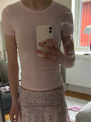 Ljusrosa tight t-shirt - Säljer en ljusrosa tight t-shirt med rund hals och korta ärmar. Toppen har en enkel och stilren design utan synliga loggor eller detaljer. Perfekt att matcha med kjol eller jeans för en clean look.