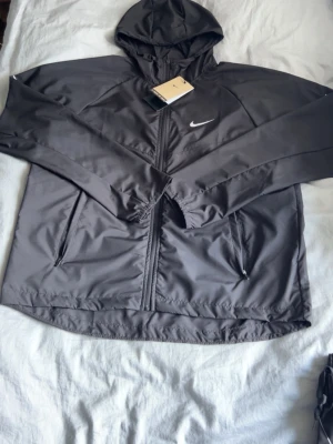 Svart Nike vindjacka med huva - Snygg svart vindjacka från Nike med huva och dragkedja framtill. Jackan har två sidofickor med dragkedja och en vit Nike-logga på bröstet. Tillverkad i lätt och vindtätt material, perfekt för blåsiga dagar. Sportig och stilren design.