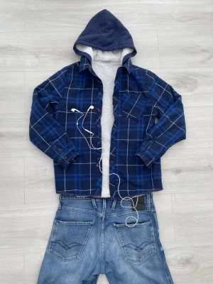 Blå rutig overshirt med huva - Snygg blå overshirt med rutigt mönster i blått och vitt. Jackan har en mjuk, ljus teddyfodrad insida och en mörkblå huva. Långa ärmar med knappar vid ärmslut och klassisk skjortkrage. Perfekt lager på lager-plagg för en avslappnad stil.