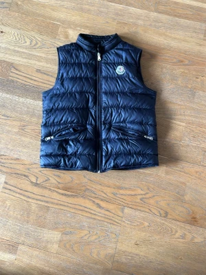 Svart dunväst från Moncler - Snygg svart dunväst från Moncler med klassisk logga på bröstet. Västen har två dragkedjeförsedda fickor framtill och stängs med dragkedja. Tillverkad i glansigt material och har en quiltad design som ger en sportig och trendig look.