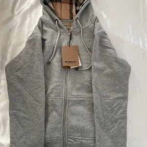 Grå hoodie från Burberry med dragkedja - Snygg grå hoodie från Burberry med klassiskt rutigt foder i huvan. Tröjan har dragkedja framtill, snörning i huvan och två fickor. Perfekt för dig som vill ha en stilren och exklusiv look med ikoniska Burberry-detaljer.
