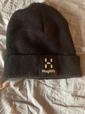 Svart mössa från Haglöfs - Svart ribbstickad mössa från Haglöfs med uppvikt kant och tydlig logotyp framtill. Klassisk och enkel design som passar till många stilar. Materialet är mjukt och bekvämt, perfekt för kalla dagar.