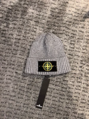 Grå ribbstickad mössa Stone Island - Säljer en grå ribbstickad mössa från Stone Island med uppvikt kant och den klassiska svarta logotyp-patchen framtill. Mössan är i mjukt material och har en clean, modern look som passar perfekt till streetwear och casual outfits. Den har aldrig varit använd, alla taggar sitter kvar och påse fås med.