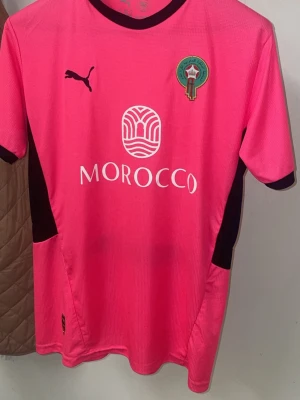 Marocko landslagströja från Puma - Snygg rosa Marocko landslagströja från Puma med svart krage och detaljer. Tröjan har tryckt landslagsemblem på bröstet och Puma-logga. Tillverkad i ett lätt och funktionellt syntetmaterial, perfekt för dig som gillar sportig stil.
