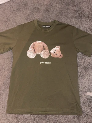 Palm Angels grön t-shirt med björntryck - Säljer en grön t-shirt från Palm Angels med ikoniskt björntryck på bröstet och logga under. T-shirten har rund hals och korta ärmar. Materialet är mjuk bomull som känns skön mot huden. Perfekt för dig som gillar streetwear och vill sticka ut.