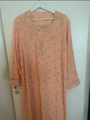 Brokadbroderad persikofärgad klänning - Persikofärgad långklänning med vackra broderier och pärldetaljer runt v-ringningen. Klänningen har diskret blommönster, trekvartsärm och är tillverkad i ett lätt tyg som känns luftigt. Perfekt för dig som gillar eleganta detaljer och unik design.