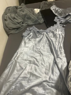 Silvrigt satinsovset med spetsdetaljer - Snyggt pyjamasset i silvrig satin med spetsdetaljer. Setet består av ett linne med spetskant vid urringningen och ett par matchande shorts med spetsmönster vid bensluten. Perfekt för dig som gillar en lyxig och mjuk känsla på natten.