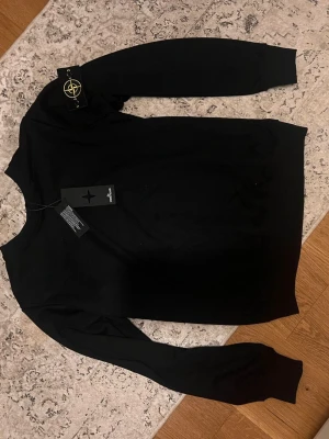 Svart tröja från Stone Island - Svart tröja från Stone Island med rund hals och lång ärm. Tröjan har den klassiska logotypen på vänster ärm och är tillverkad i mjukt material. Enkel och stilren design som lyfter din outfit.