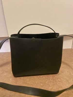 Svart axelväska i läderlook - Snygg svart axelväska med stilren, fyrkantig form och bred axelrem. Väskan har även ett praktiskt handtag upptill och är gjord i ett material som liknar läder. Perfekt för dig som gillar minimalistisk design och vill ha plats för det viktigaste.