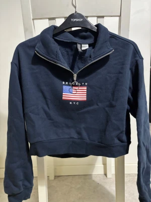 Croppad mörkblå tröja med litet tryck  - Mörkblå tröja med litet tryck framtill och en dragkedja upptill.