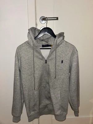 Grå zip hoodie från Polo Ralph Lauren - Snygg grå hoodie med dragkedja från Polo Ralph Lauren. Klassisk design med huva och dragsnören, samt den ikoniska lilla loggan broderad på bröstet. Tillverkad i mjukt bomullsmaterial som är skönt att ha på sig. Perfekt till chill dagar eller när du vill ha en clean look.