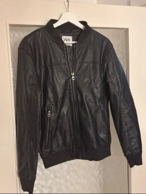 Svart skinnjacka från Zara - Snygg svart skinnjacka från Zara med dragkedja framtill och två fickor med dragkedja. Jackan har ribbade muddar vid ärmslut och nederkant samt en klassisk bomberkrage. Perfekt för dig som vill ha en stilren och tidlös look.