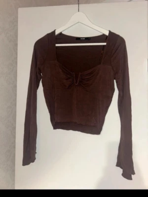 Brun croppad topp med rynkdetalj - Snygg brun croppad topp från Bik Bok med långa ärmar och rynkdetalj framtill. Toppen har en fyrkantig ringning och är tillverkad i ett mjukt, stretchigt material som sitter bekvämt. Perfekt att styla med höga jeans eller kjol.
