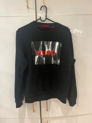Svart sweatshirt från HUGO med tryck - Säljer en svart sweatshirt från HUGO med ett stort grafiskt tryck i svart, vitt och grått på bröstet samt röd HUGO-logga. Tröjan har rund halsringning och långa ärmar. Perfekt för dig som gillar streetwear och vill sticka ut. Som passar FOLK med storlek xs, skriv för mer bilder