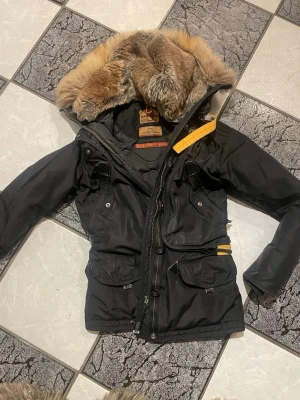 Svart parkas från Parajumpers XS/S - Snygg svart parkas från Parajumpers med stor luva fodrad i beige fuskpäls och detaljer i gult. Jackan har flera fickor framtill, dragkedja och knappar samt justerbar midja. Perfekt för kalla dagar och riktigt varm. Jackan är i fint skick endast använd 1 säsong och ett fåtal gånger, bilderna ger inte jackan rättvisa den är verkligen i nyskick. Den inköptes på Emporia för 4 år sedan men använd under en säsong sen har den varit inhängd i garderoben. Lågt utropspris! 