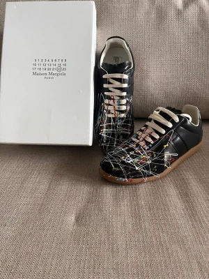 Maison Margiela svart sneakers konstprint - Maison Margiela sneakers i svart läder med färgstänk i vitt, rött, gult och blått över hela ovandelen. Klassisk låg modell med vita snören och brun gummisula. Unik design som sticker ut och passar perfekt för dig som gillar streetwear och konstnärliga detaljer.
