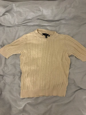 Beige stickad topp från Vero Moda - Snygg beige stickad topp från Vero Moda med korta ärmar och rund hals. Tröjan har ett diskret hålmönster och ribbade kanter vid ärmslut och nederkant. Perfekt till jeans eller kjol för en clean look.