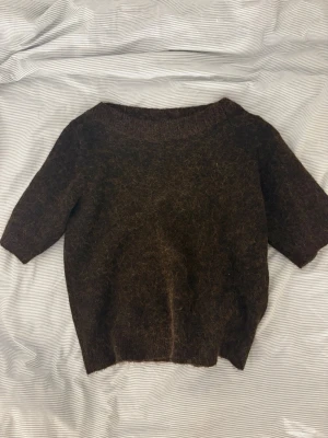 Brun stickad tröja Gina Tricot - Mysig brun stickad tröja från Gina Tricot med korta ärmar och bred halsringning. Tröjan har en fluffig, lite luddig yta och ribbstickade detaljer vid hals och nederkant. Perfekt för lager på lager och en avslappnad look.