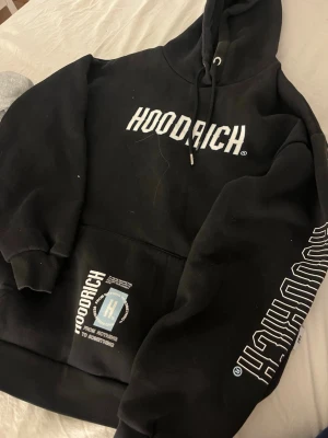 Svart HOODRICH hoodie med tryck - Svart hoodie från HOODRICH med stor vit logga på bröstet och ärmen samt tryck på fickan. Klassisk känguruficka och justerbar huva med snören. Tillverkad i mjukt bomullsmaterial för en skön och avslappnad look.