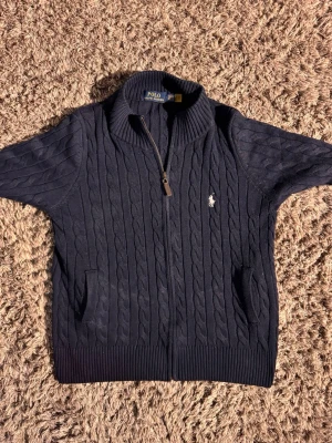 Mörkblå kabelstickad zip -tröja Polo🙌🤩 - Mörkblå kabelstickad tröja från Polo Ralph Lauren med dragkedja framtill, ribbad krage och två fickor. Ikonisk broderad logga på bröstet. Perfekt för dig som gillar klassisk och stilren design.🙌🤩