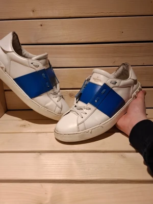 Valentino Open vita sneakers med blå detalj i storlek 43 - Snygga vit blåa Valentino Opens från Valentino Garavani med en bred blå rem över mitten. Skorna har klassisk låg profil, vita snören och diskret logga på plösen. Tillverkade i läder med stilren design som sticker ut med den blåa detaljen. De är i riktigt bra skick och väl omhändetagna, de är i storlek 43. Bara att fråga om ni vill ha mer bilder eller diskutera priset, Skickar samt svarar väldigt fort
