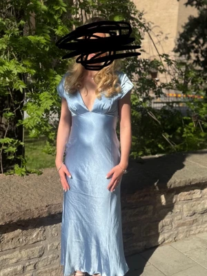 Ljusblå v-ringad klänning i satin - Ljusblå lång klänning med djup v-ringning och korta ärmar. Klänningen har en elegant, flowy passform och är tillverkad i glansigt satinmaterial som ger en lyxig känsla. Perfekt för dig som vill sticka ut med en stilren look.