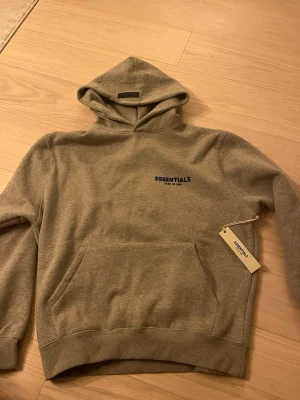 Grå hoodie från Essentials Fear of God - Snygg grå hoodie från Essentials Fear of God med diskret logga på bröstet. Klassisk passform med huva och stor magficka. Tillverkad i mjukt bomullsmaterial som känns riktigt skönt mot huden. Perfekt för en avslappnad och trendig look.