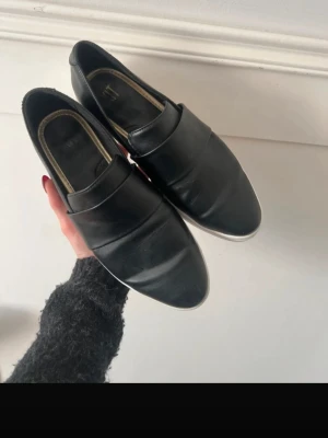 Svarta loafers från H&M - Säljer ett par stilrena svarta loafers från H&M med klassisk design och rundad tå. Skorna har en bred rem över foten och vit sula med mönster undertill. Materialet är slätt och ger en snygg look som funkar till många outfits.
