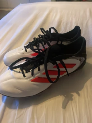 Adidas vita och röda fotbollsskor - Snygga fotbollsskor från Adidas med vit ovandel, röda detaljer och svarta snören. Skorna har klassisk låg profil och mönstrad sula med dobbar för bra grepp på planen. Materialet är syntetiskt och designen är modern med sportiga linjer.