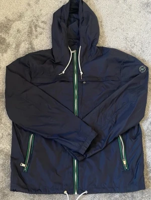 Marinblå vindjacka från Polo Ralph Lauren - Snygg marinblå vindjacka från Polo Ralph Lauren med huva och vita dragskor. Jackan har gröna och vita detaljer på dragkedjorna samt två sidofickor med dragkedja. Liten Polo-logga på ärmen. Tillverkad i lätt, vindtätt material.