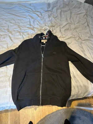 Svart zip-hoodie från Burberry - Säljer en svart hoodie med dragkedja från Burberry. Hoodien har klassiskt rutigt foder i huvan i svart, vitt och rött. Två stora fickor framtill och ribbade muddar vid ärmslut och nederkant. Perfekt för dig som gillar stilrena och exklusiva plagg.OBS inga snören. Diskutera pris.