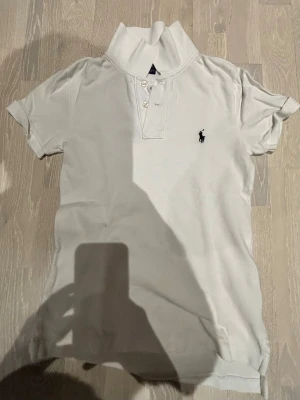 Vit pikétröja från Ralph Lauren - Klassisk vit pikétröja från Ralph Lauren med korta ärmar och den ikoniska broderade loggan på bröstet. Tröjan har krage, knappar framtill och är tillverkad i mjuk bomull. Perfekt till jeans eller shorts för en clean look.