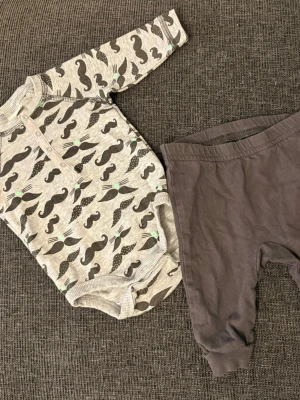 body med mustaschmönster & byxa - Säljer ett set med en långärmad body med mustascher och små gröna detaljer samt matchande mörkgrå byxa. Bodyn har tryckknappar framtill och i grenen för enkel påklädning. Body från Lindex byxor lupilu 