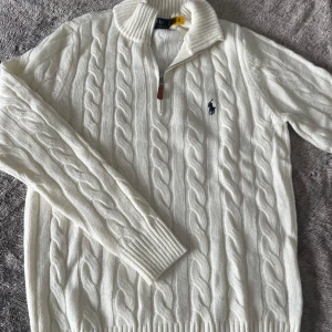 Vit kabelstickad tröja Polo Ralph Lauren - Kabelstickad vit tröja från Polo Ralph Lauren med halv dragkedja och ribbad krage. Tröjan har långa ärmar, ribbade muddar och den klassiska mörkblå logotypen broderad på bröstet. Perfekt för en stilren och klassisk look. Bra skick