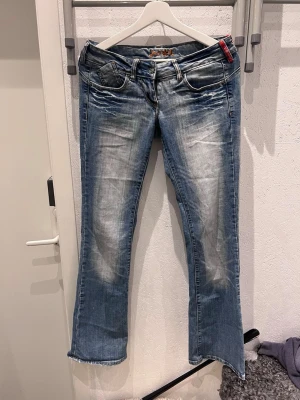 Blå bootcut jeans med brodyr - Säljer ett par blå bootcut jeans med slitningar och cool brodyr i orange och blått på bakfickorna. Jeansen har låg midja, fem fickor och detaljer i kontrastsömmar. Perfekta för dig som gillar en avslappnad och trendig stil. De är små i storleken! 