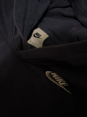 Mörkblå hoodie från Nike - Snygg mörkblå hoodie från Nike med klassisk broderad logga på bröstet. Hoodien har en stor huva och magficka framtill. Tillverkad i mjukt bomullsmaterial som är skönt att ha på sig. Perfekt för en avslappnad och sportig stil.