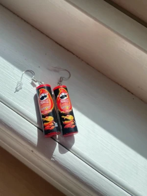 Pringles örhängen svart/röd - Unika örhängen formade som små Pringles-rör i svart och rött med Super Hot Chili & Cheese-design. Örhängena har krokar i metall och detaljerade tryck som liknar riktiga chipstuber. Perfekt för dig som gillar statement-accessoarer och vill sticka ut.