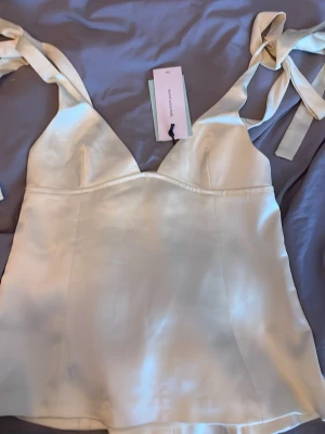 Champagnefärgad satinlinnetopp med knyt - Säljer en champagnefärgad linnetopp i satin med breda axelband som knyts till rosetter. Toppen har en lätt v-ringad form och slät finish som ger en lyxig känsla. Perfekt för dig som vill ha en elegant och trendig topp.