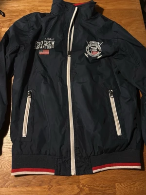 Marinblå vindjacka Polo Crew - Snygg marinblå vindjacka från Vinson Polo Club med vita dragkedjor, broderade detaljer och amerikansk flagga på bröstet. Jackan har ribbade muddar med röd och vit rand, hög krage och två fickor framtill. Perfekt för våren. 