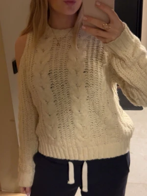 Vit stickad tröja med cold shoulder - Säljer en krämvit stickad tröja med flätmönster och trendiga cold shoulder-detaljer. Tröjan har rund halsringning, långa ärmar och ribbade muddar. Perfekt för dig som vill ha en mysig men ändå unik look.